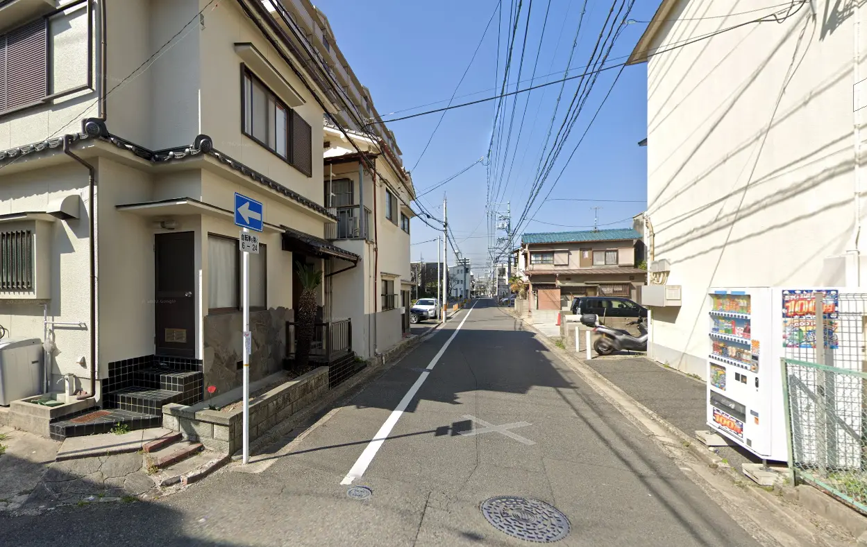 狭い住宅街の一方通行の路地、日中の晴れた日、左手には自動販売機が見え、右手には民家が立ち並ぶ。