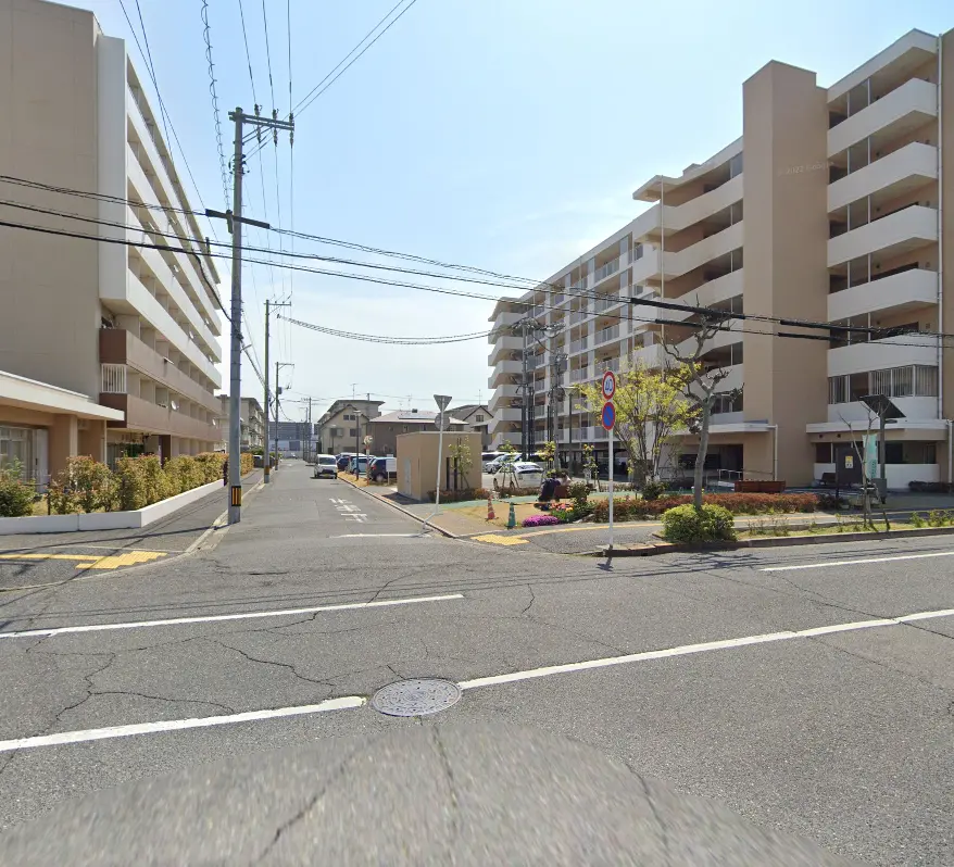 高層マンションが並ぶ住宅街の交差点。両側には整然とした植栽や駐車場があり、落ち着いた雰囲気が漂っています。