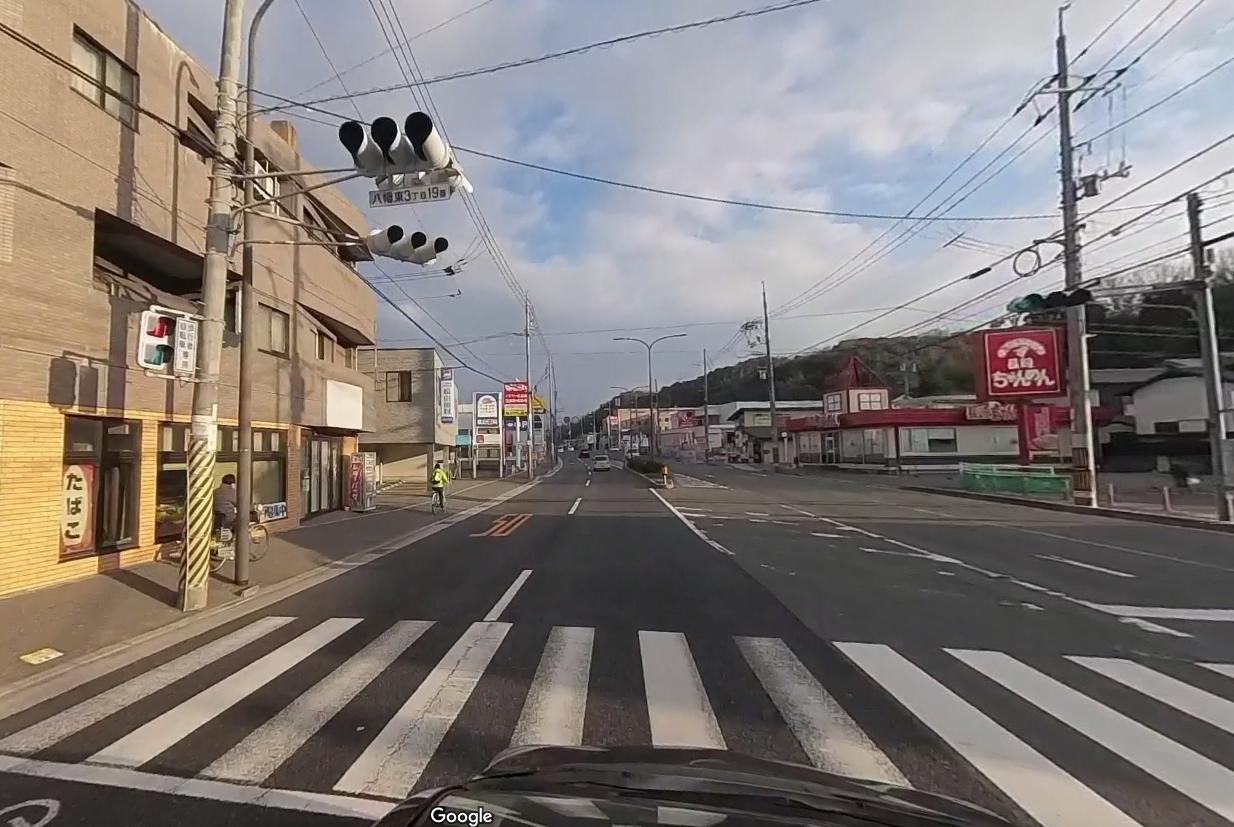 信号機と横断歩道がある交差点の風景。車道の両側には商業施設や看板が並んでおり、道路沿いを自転車に乗る人物が見られる。