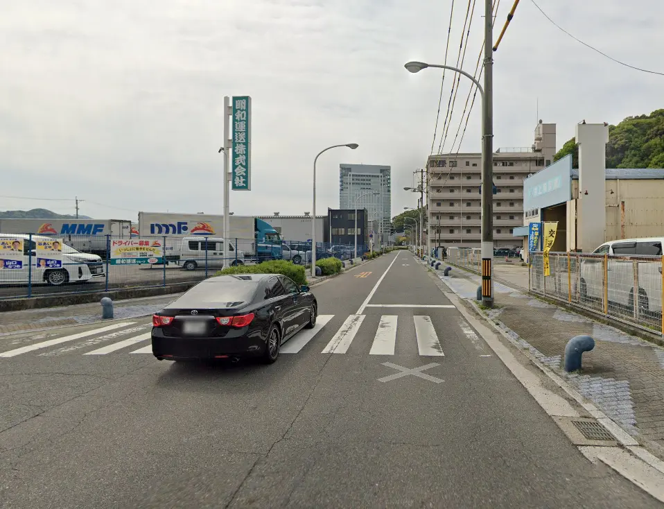 工場や物流施設が並ぶ道路に、黒い車が進んでいる風景。道路の両側には倉庫やビルが立ち並び、看板やフェンスが見られます。