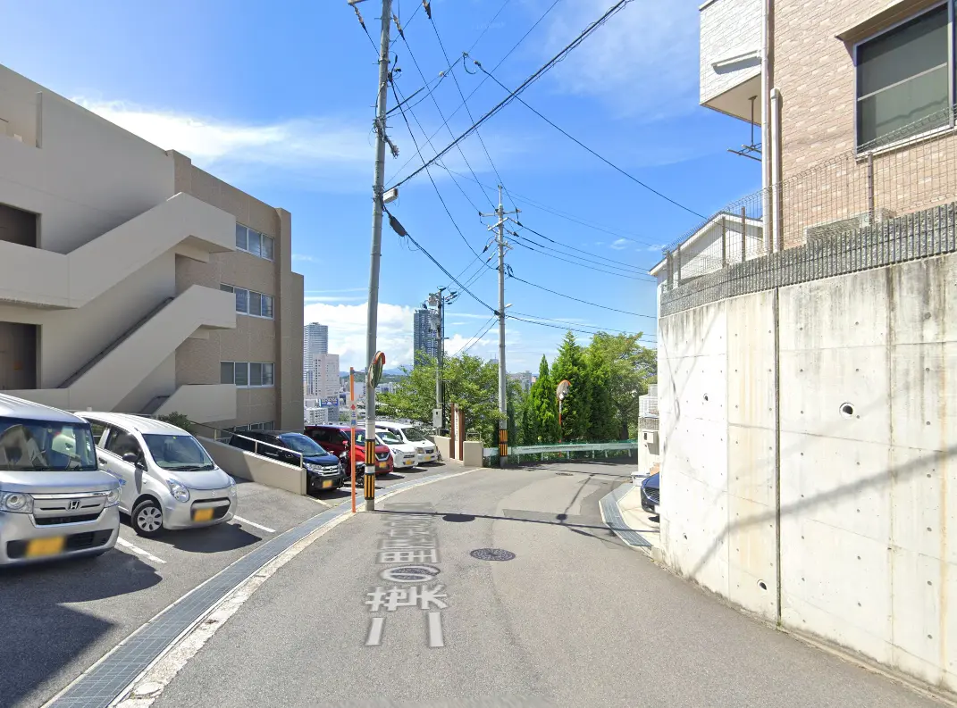 坂道に沿った駐車場と建物の風景。晴れた空が広がり、遠くにビル群が見えます。