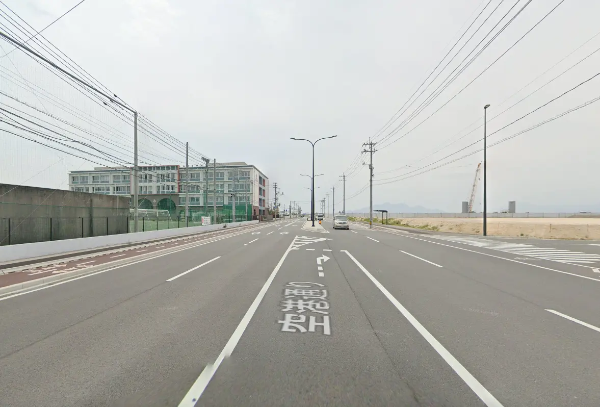 広々とした道路に面する建物と電線が交差する風景。左側には学校のような建物とサッカーグラウンドが見え、右側には空き地が広がり、遠くに山々が見える。