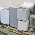 数台の古い家電製品が道路沿いに置かれている。冷蔵庫、洗濯機、エアコン、テレビなどが並んでいる。