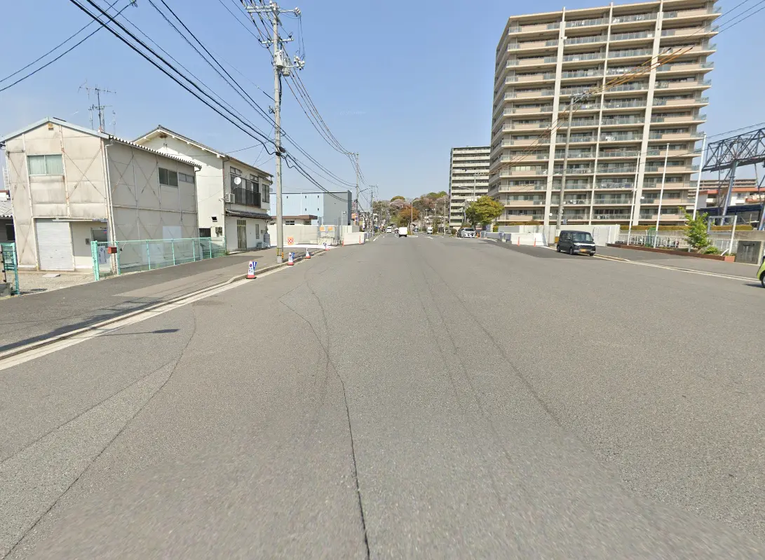 静かな住宅街の道路。左側に古い建物、右側には高層マンションが並んでおり、広々とした道路がまっすぐに続いています。